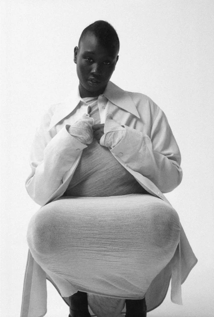 Woman with shaved head in light grey dress and open collared shirt in black and white studio shot with white backdrop, neutral expression.