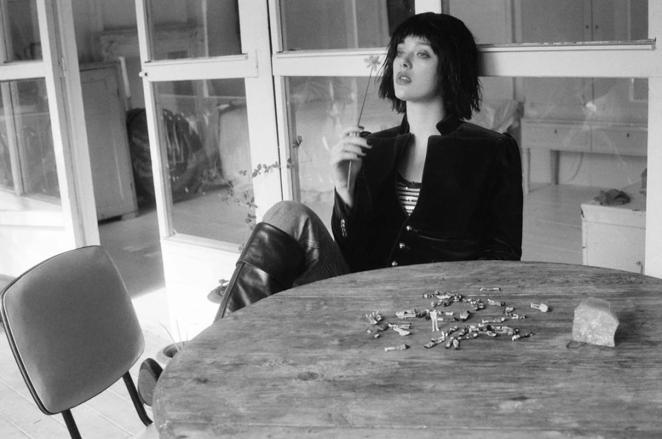 Black and white portrait of a young adult female seated at a table with a flower, wearing a velvet jacket, and looking away.