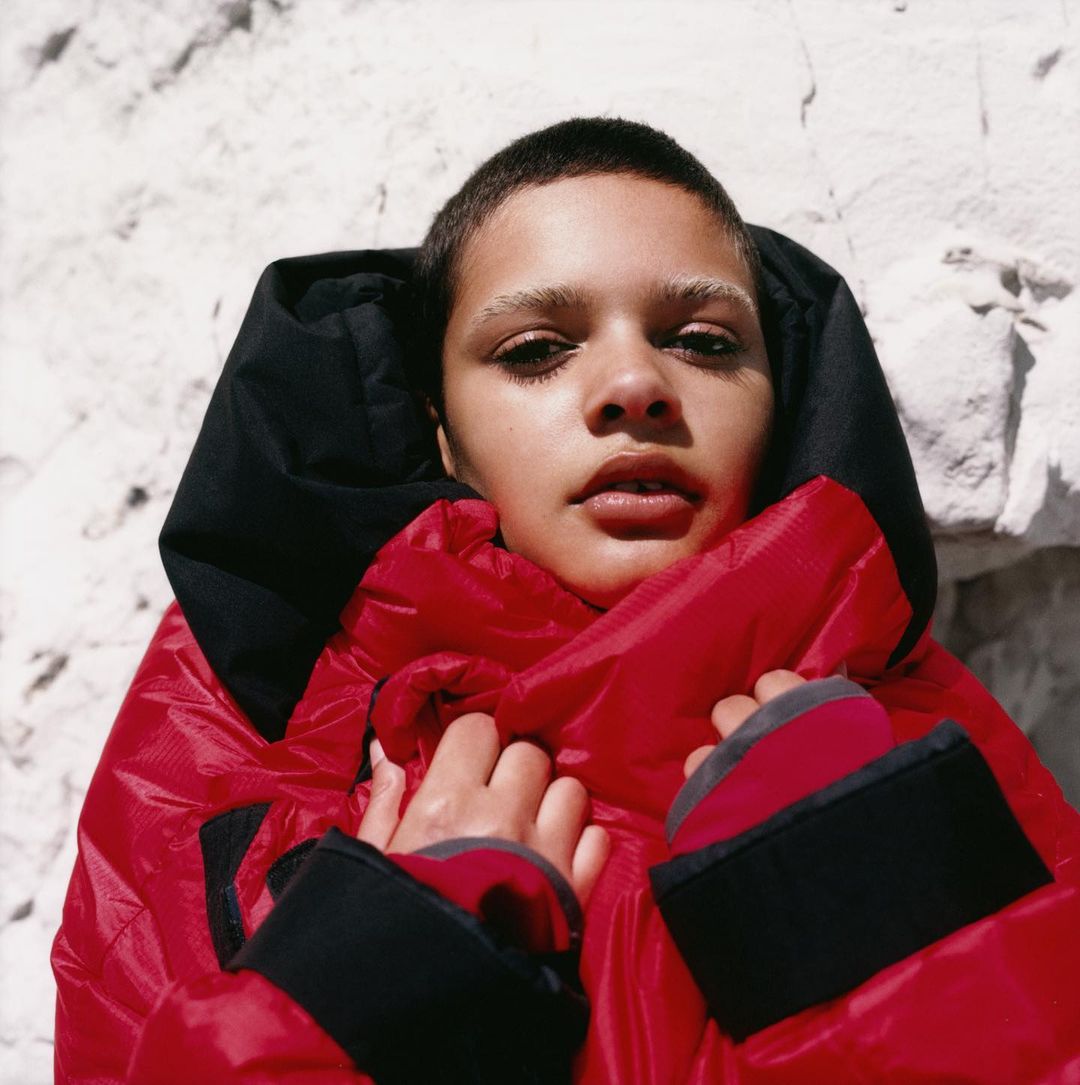 A young adult woman with short hair poses in a bright red puffer coat with a black hood against a textured white background.