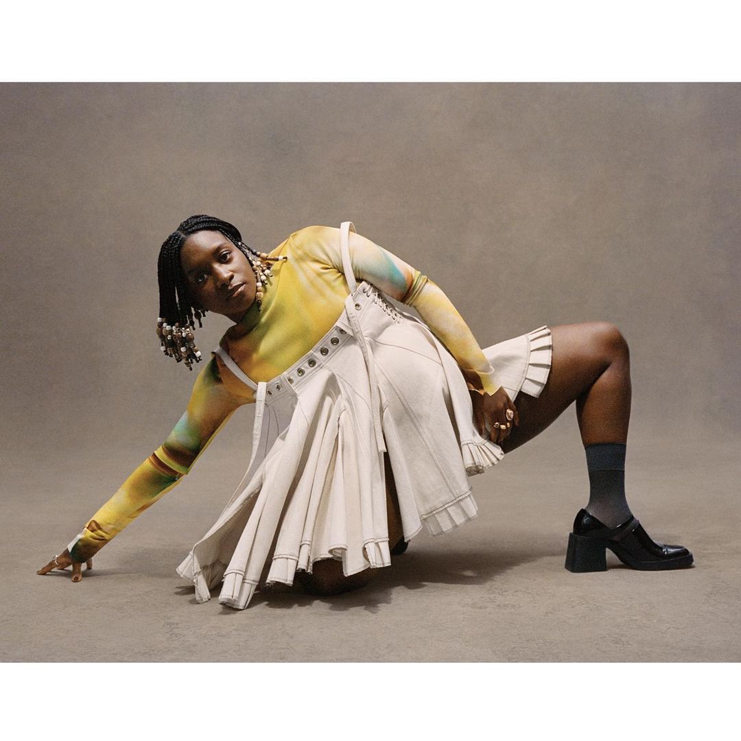 A Black woman posing in a yellow tie dye top and cream pleated skirt against a neutral studio backdrop.