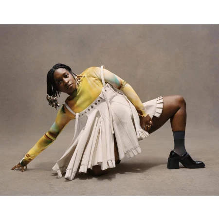 A Black woman posing in a yellow tie dye top and cream pleated skirt against a neutral studio backdrop.