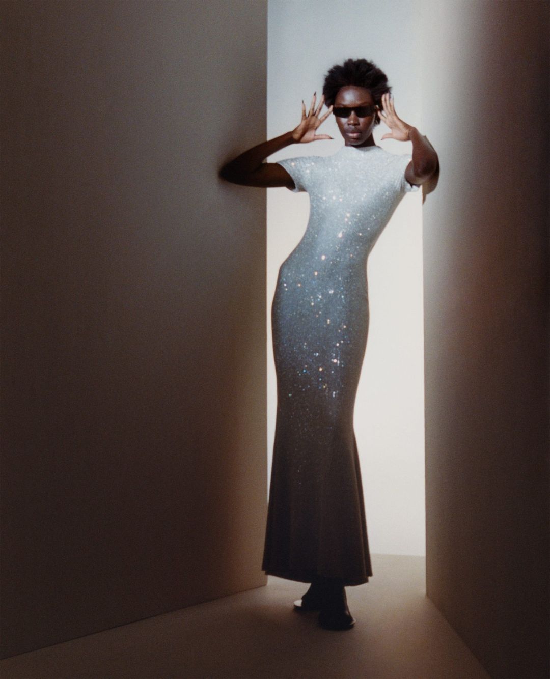 Elegant black woman in a shimmering silver gown poses in a photo studio, showcasing a glamorous fashion aesthetic with soft lighting.