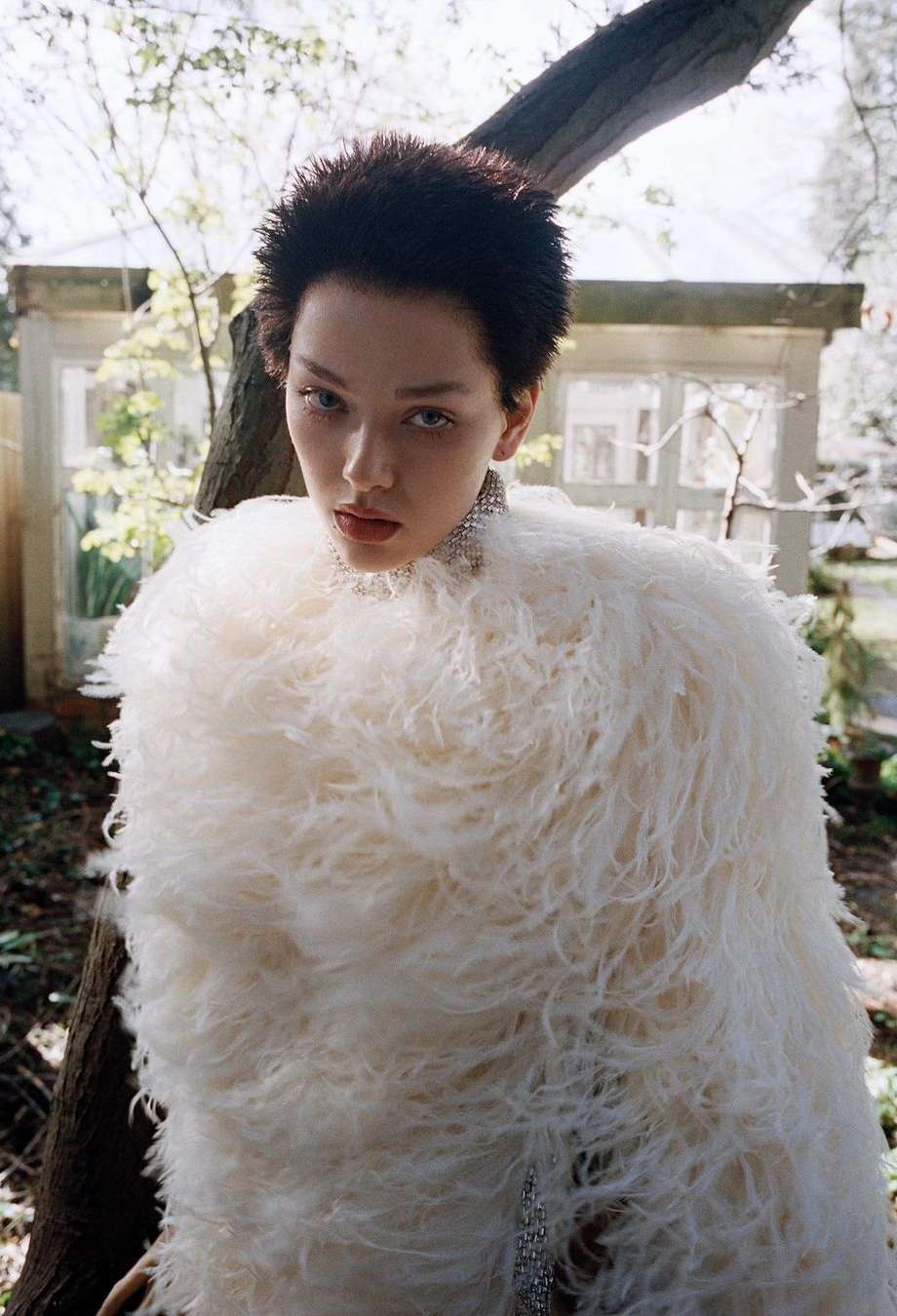 Woman in a fluffy white jacket stands outdoors, looking at the camera with trees and a building in the blurred background.