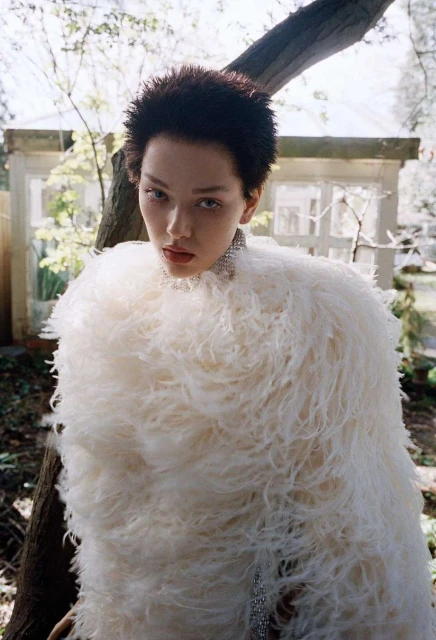 Woman in a fluffy white jacket stands outdoors, looking at the camera with trees and a building in the blurred background.
