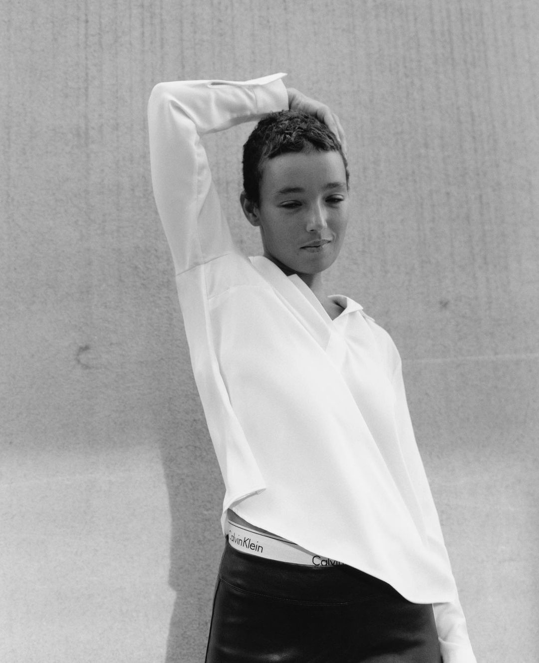 Portrait of a slender young woman in white shirt and black leather pants poses against a concrete wall in a minimalist, black and white style.