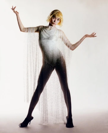 Woman with short blonde hair wearing a sheer sparkly cape over a white leotard and black heeled mules against a plain light background