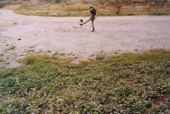 A woman in patterned clothing kicks a hat up in the air on a gravel road, set against a backdrop of grassy ground cover and distant vegetation.
