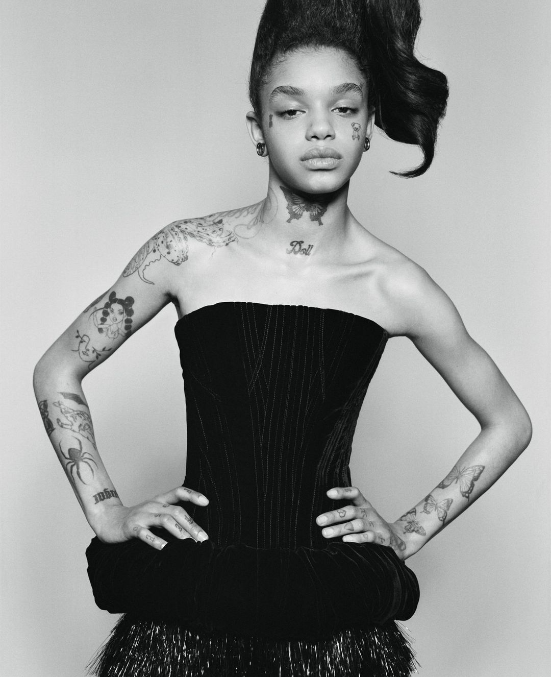 Monochrome portrait of a young adult woman with a high hairstyle, wearing a black dress with shoulder tattoos in a light studio.