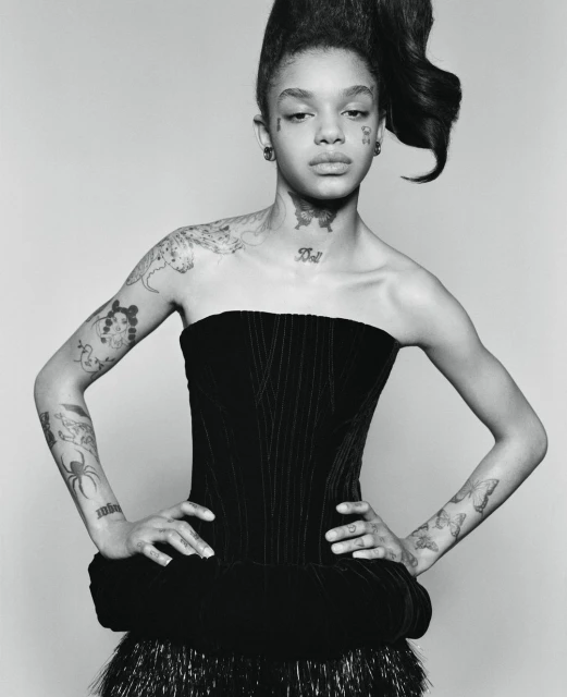 Monochrome portrait of a young adult woman with a high hairstyle, wearing a black dress with shoulder tattoos in a light studio.