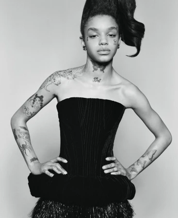 Monochrome portrait of a young adult woman with a high hairstyle, wearing a black dress with shoulder tattoos in a light studio.