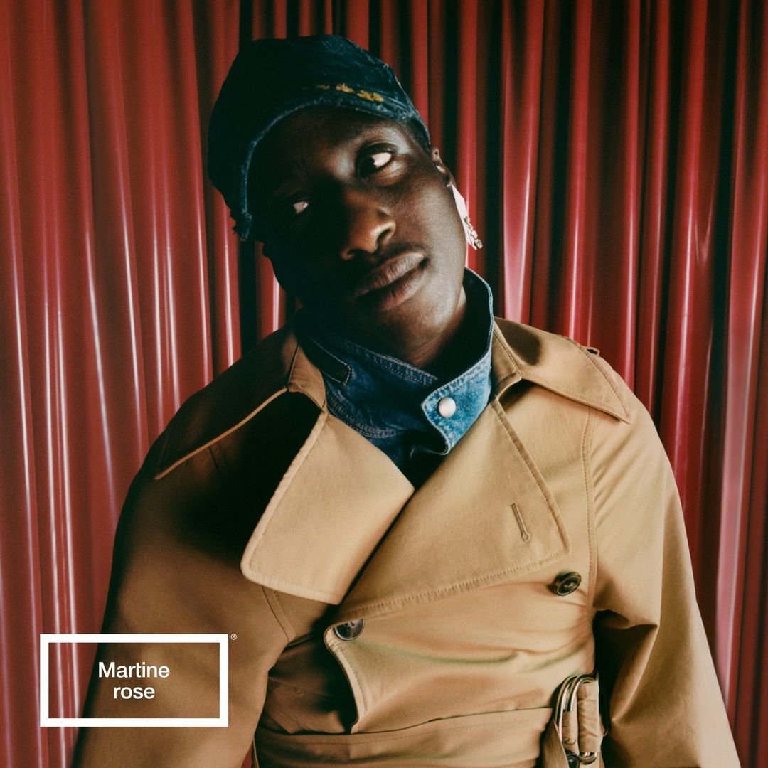 Close-up studio shot of an adult Black male model with a denim shirt, beige trench coat and patterned cap against a red curtain backdrop.