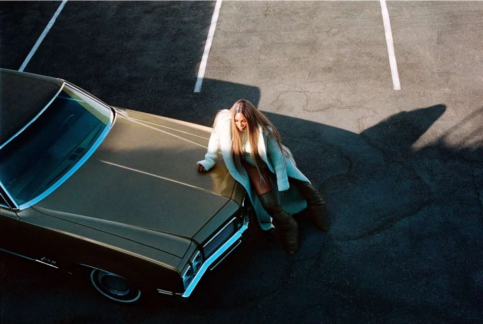 Woman in coat leaning on a vintage car in a parking lot with painted stripes, bathed in warm light. Fashion and automotive theme.