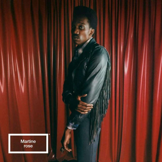 Stylish Black male model poses in a black fringed jacket against a red draped background; fashion-forward and captivating.