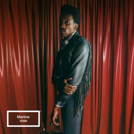 Stylish Black male model poses in a black fringed jacket against a red draped background; fashion-forward and captivating.