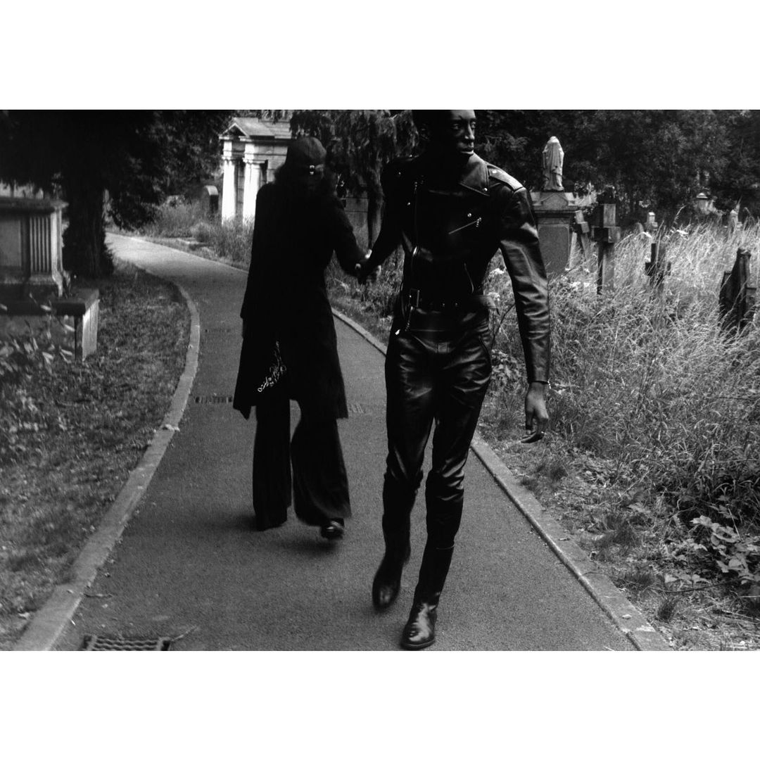 Two figures in dark clothing hold hands walking through a cemetery. Tall grass, trees, and gravestones are visible in a black and white outdoor scene.