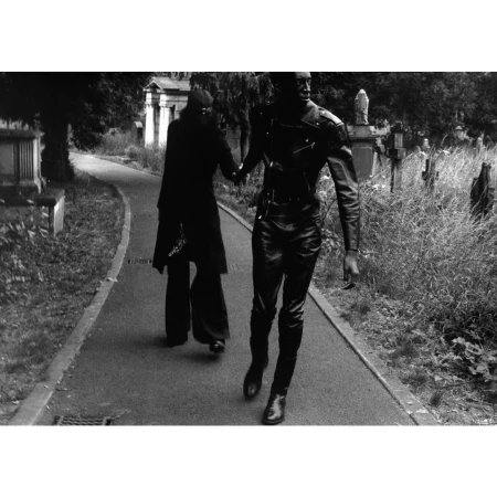 Two figures in dark clothing hold hands walking through a cemetery. Tall grass, trees, and gravestones are visible in a black and white outdoor scene.