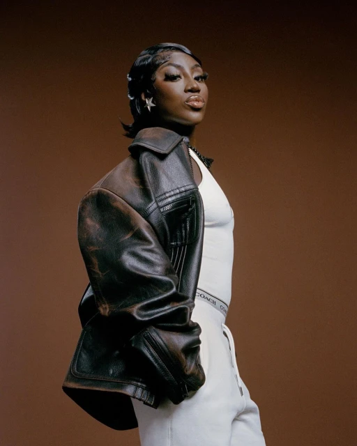 Portrait of a Black woman in a white outfit with a brown leather jacket and star earrings against a solid brown backdrop.