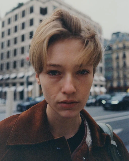 Close-up portrait of a fair-skinned young adult female with short blonde hair wearing a brown jacket on a city street.