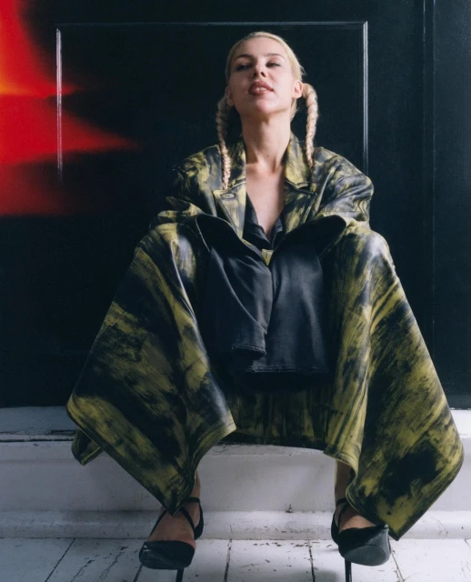 Adult woman in a bold print coat poses on a step in high heels, with pigtails and a serious expression against a dark backdrop.