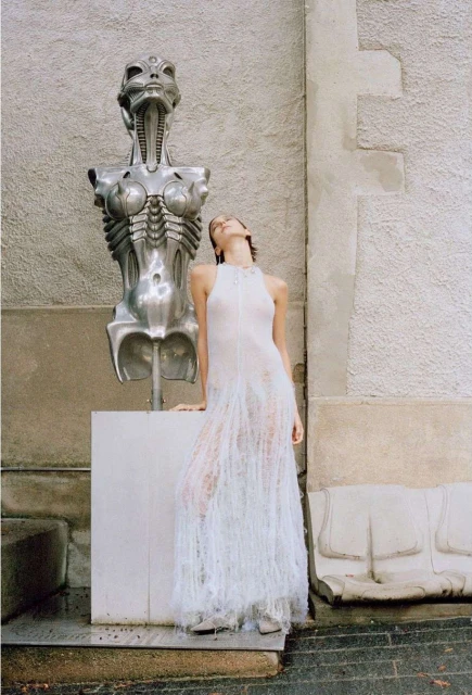 Woman in sheer dress poses next to alien-like sculpture in daylight. The statue's silver, metallic figure contrasts with the dress.