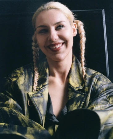Close-up portrait of a smiling woman with blonde braided pigtails wearing a patterned leather jacket by Jessica Madavo.
