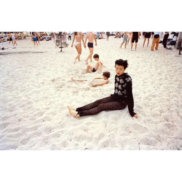 Woman sits on the sand at a crowded beach with children playing and other people enjoying the sun and surf in the background.