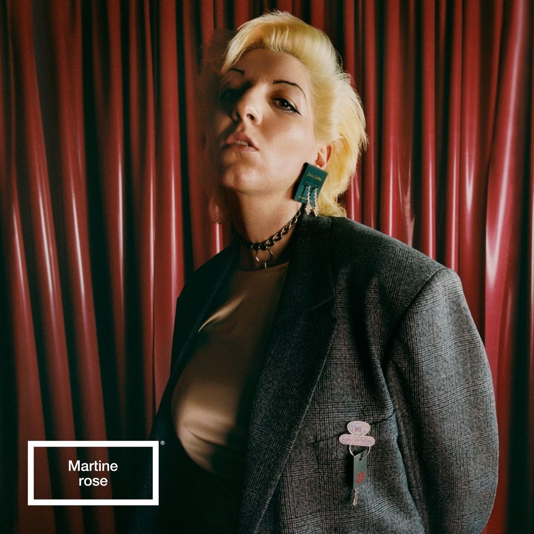 Stylish woman with blonde hair wearing a grey blazer, chunky choker, and statement earrings poses in front of a red curtain.