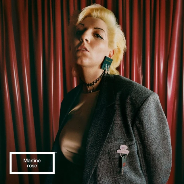 Stylish woman with blonde hair wearing a grey blazer, chunky choker, and statement earrings poses in front of a red curtain.