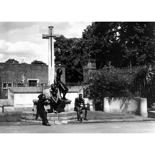 Black and white outdoor shot of a group of four adults posing near a stone cross monument with trees and a building in the background.