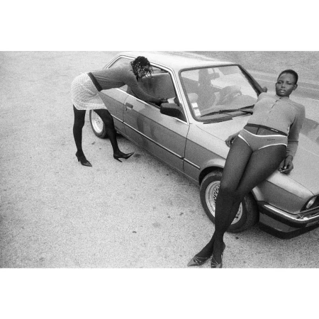 Two women posing with a vintage car in a grainy black and white photograph by Jessica Madavo
