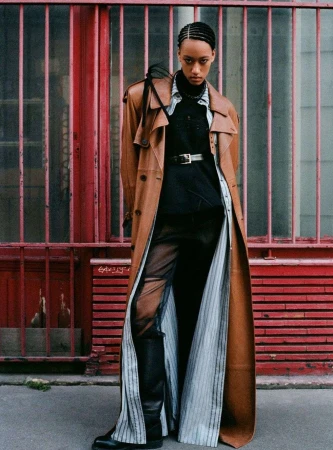 Model wearing a long brown leather coat and striped tunic standing against a red metal grating in London