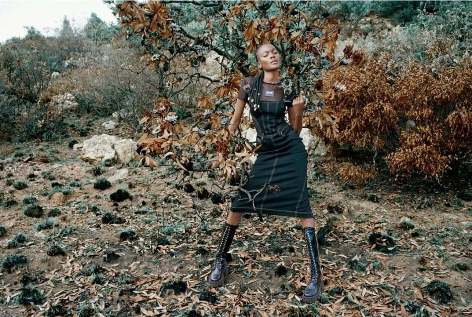 Fashionable adult woman poses in nature wearing a dark denim dress and knee-high boots, surrounded by dry leaves and trees.