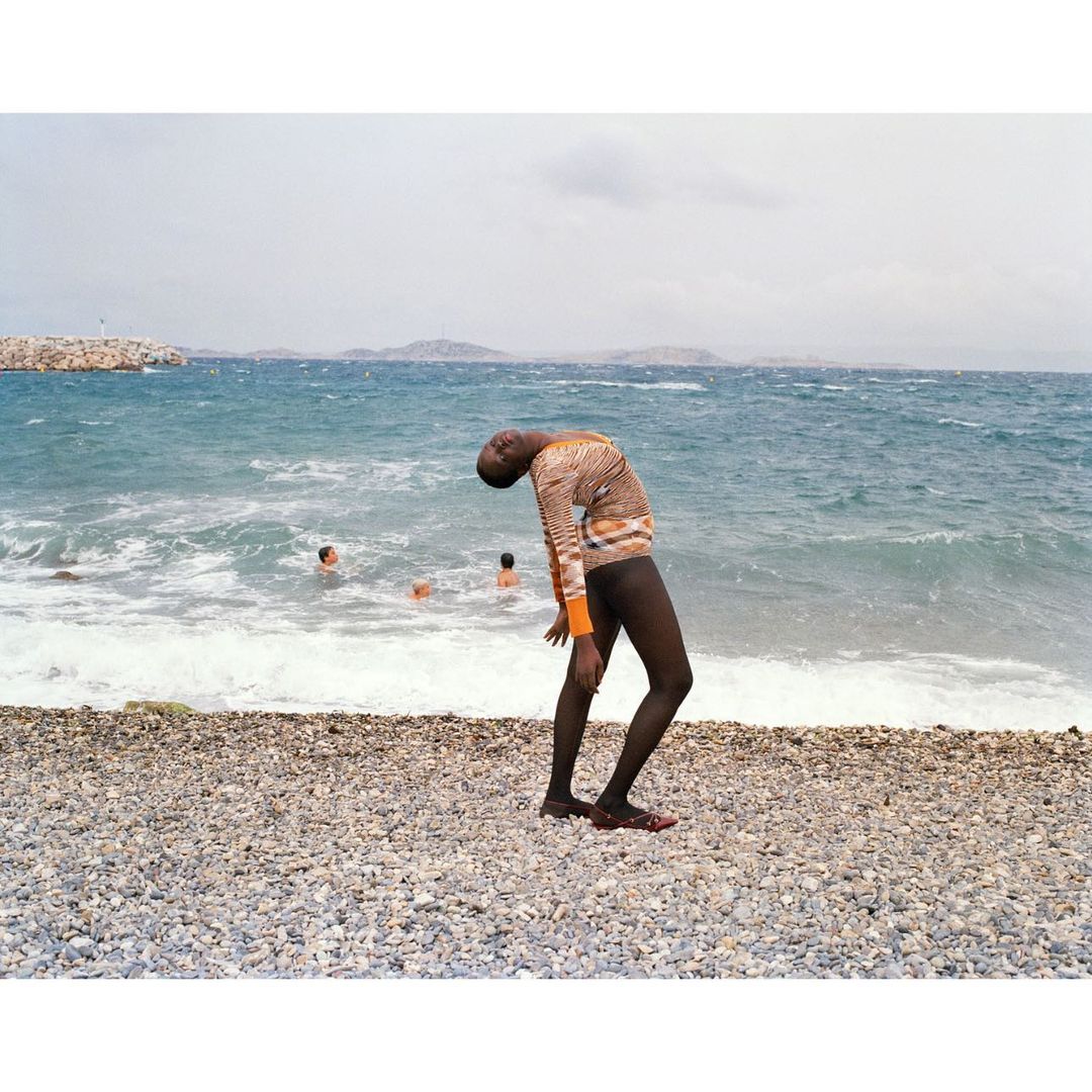 Adult standing on pebble beach with arched back, ocean waves crashing, and people swimming in background. Sunny day, open sky.