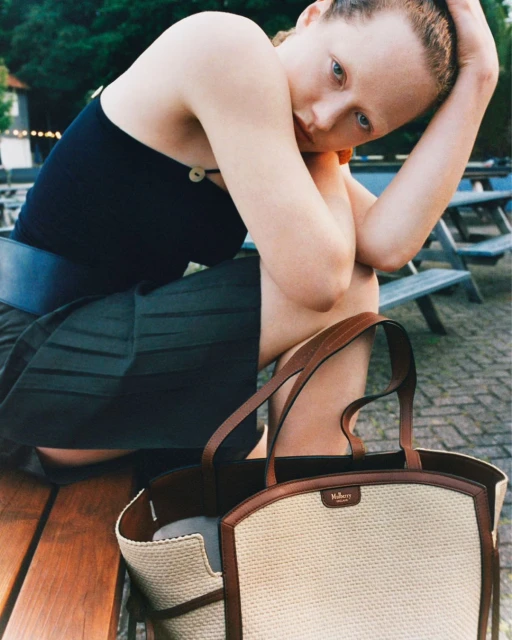 Young woman with a beige woven tote bag, resting on a wooden bench in an outdoor setting, wearing a dark top and olive skirt.