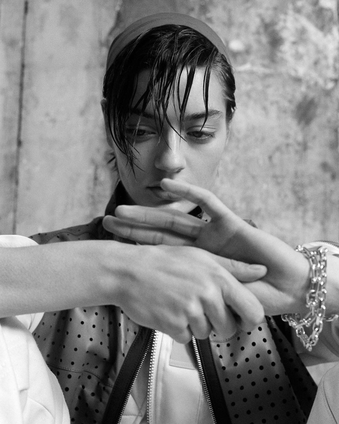Black and white portrait of a young adult woman with a headband, hands together, wearing a jacket, and bracelet against a textured wall.
