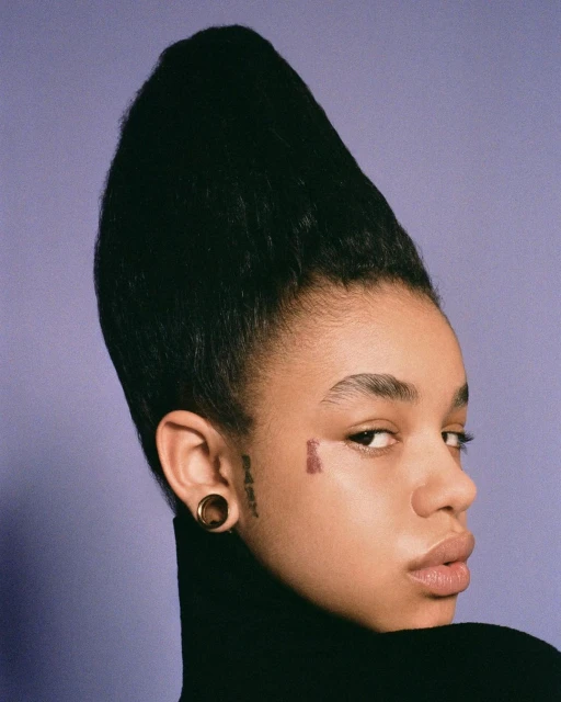 Portrait of an African-American woman with a tall, cone-shaped hairstyle, wearing a black turtleneck against a solid light purple backdrop.