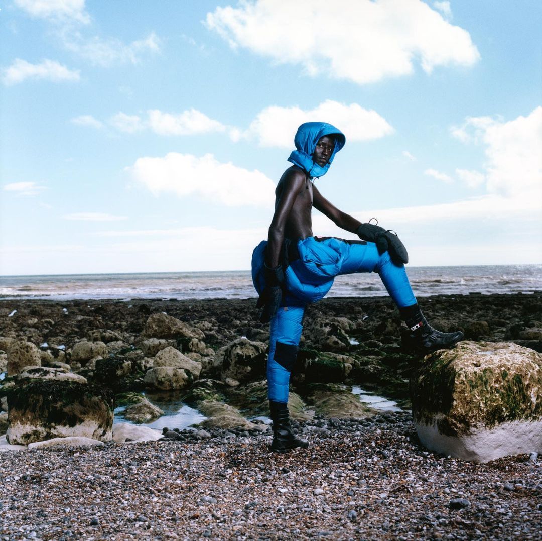 Fashion model with dark skin poses on a pebble beach wearing a blue head covering and matching trousers, standing on a rock formation.