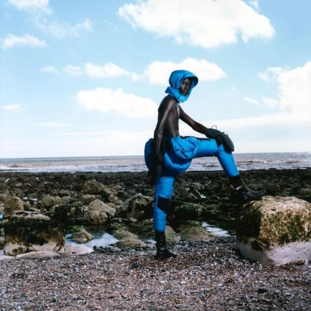 Model wearing bright blue padded puffer pants and matching hood posing on a rocky coastline by the ocean.