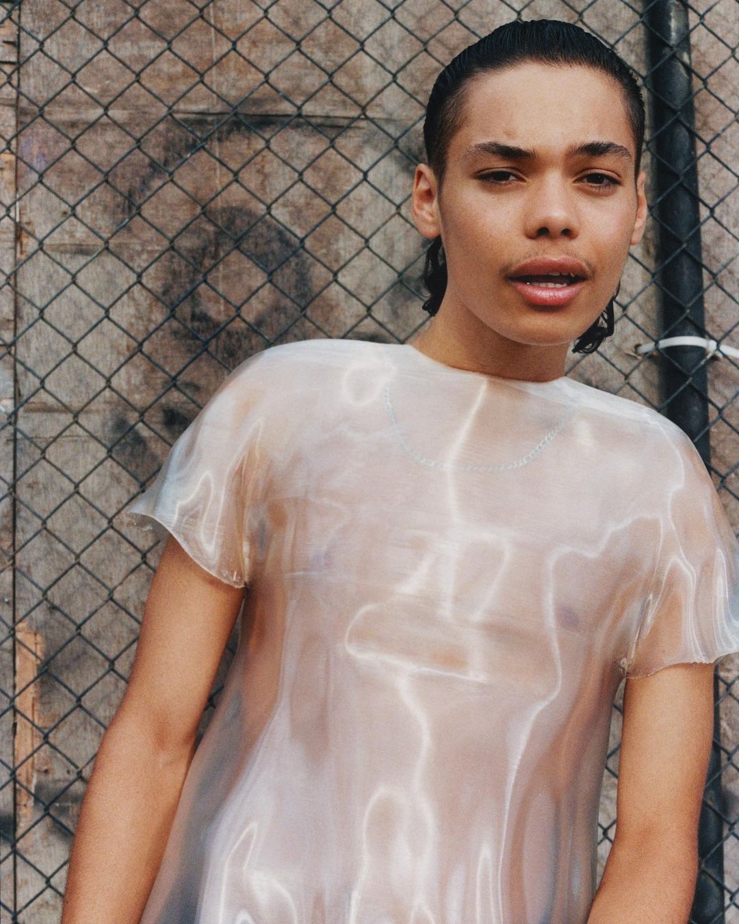 Portrait of a young person with short dark hair, wearing a transparent shirt, posing in front of a chain-link fence and textured wall.