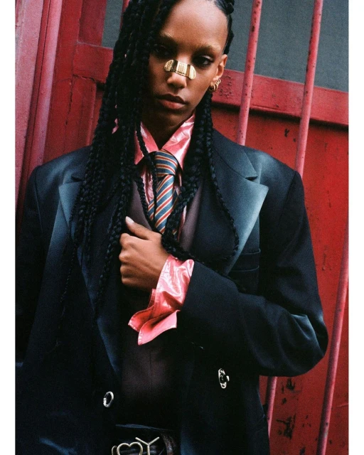 Stylish woman in oversized black suit and gold nose cuff posing against a red metal door. Fashion-forward editorial shot with dramatic lighting.