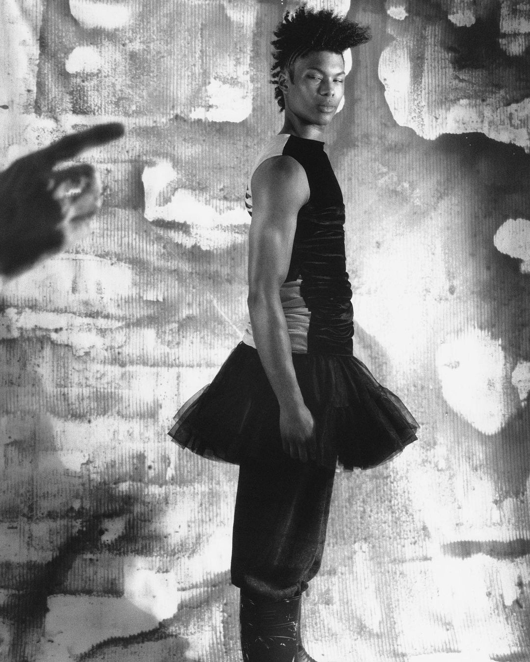 Monochromatic portrait of a person in a sleeveless top and tutu posing in front of a mottled wall with a hand pointing.