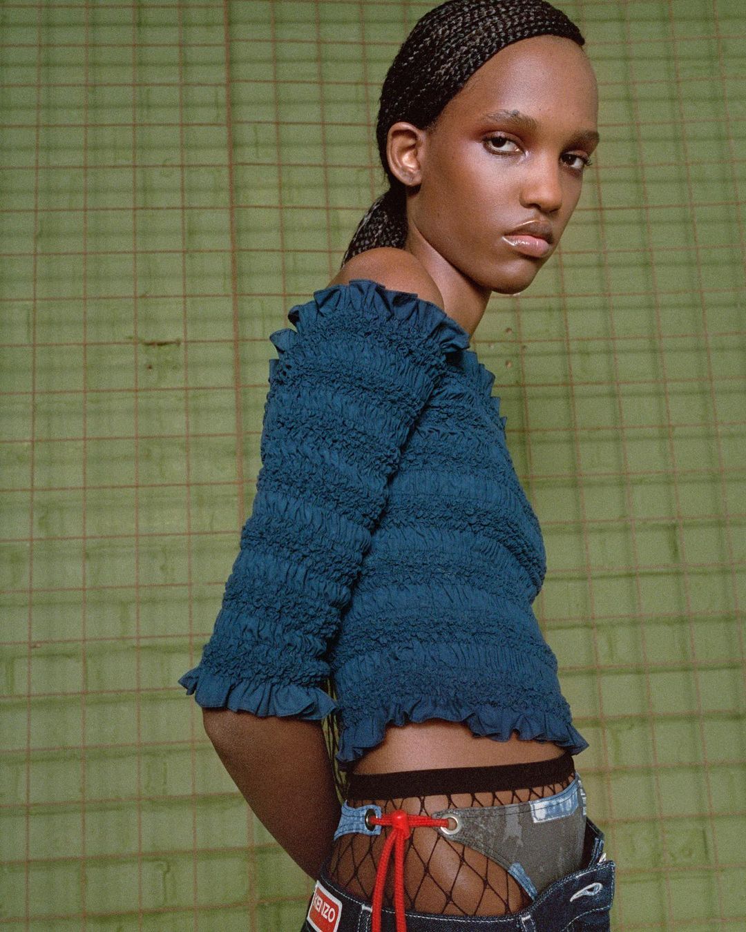 Fashion shot featuring a dark-skinned woman in a teal smocked top and deconstructed denim shorts against a textured green backdrop.