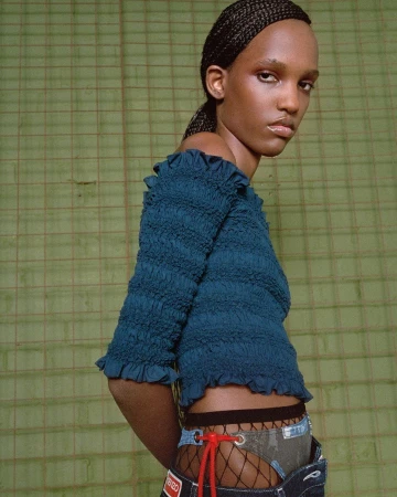 Fashion shot featuring a dark-skinned woman in a teal smocked top and deconstructed denim shorts against a textured green backdrop.