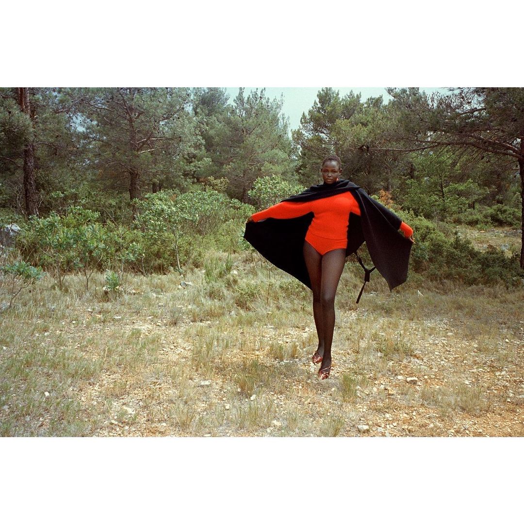 A slender Black woman wears a vibrant red bodysuit and dramatic black cape, posing in a sunlit meadow with a backdrop of lush green trees.