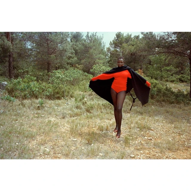 A slender Black woman wears a vibrant red bodysuit and dramatic black cape, posing in a sunlit meadow with a backdrop of lush green trees.