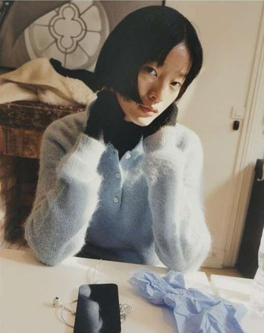 Indoor portrait of a young Asian woman with short black hair wearing a light blue cardigan, seated at a white table with a phone and gloves.
