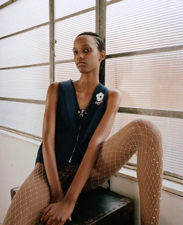 A young Black woman models a navy vest with fishnet stockings in a minimalist indoor setting by a window with natural lighting.