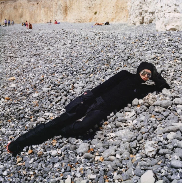 Person in a black bodysuit and sunglasses lying on a pebble beach with chalk cliffs in the background, enjoying a sunny day.