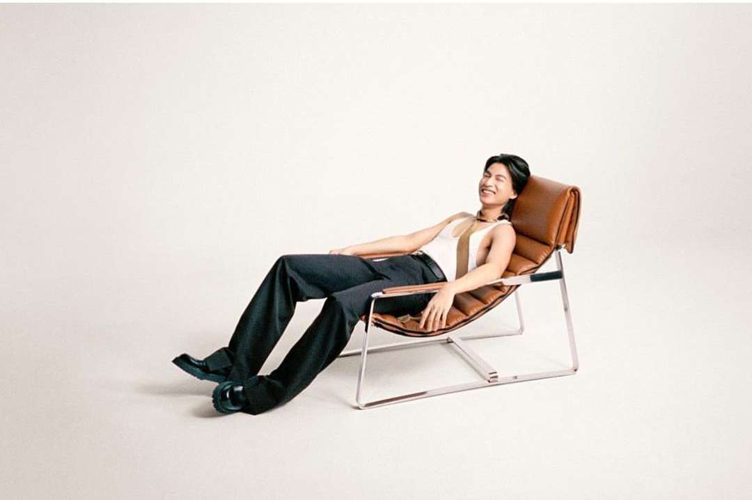 Adult male relaxes in a tan leather chair wearing a striped top and dark pants in a bright studio. Fashion portrait photography.