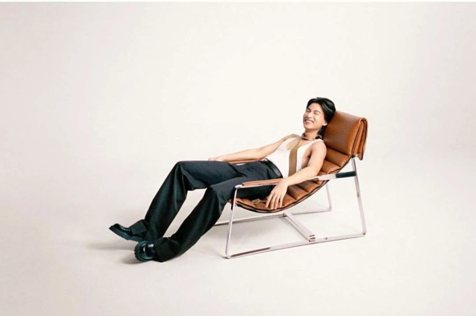 Adult male relaxes in a tan leather chair wearing a striped top and dark pants in a bright studio. Fashion portrait photography.